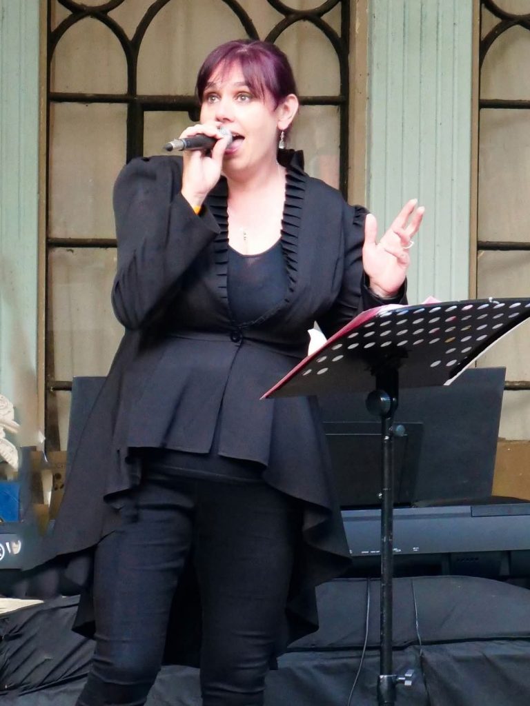 Angharad in a black frock coat and black trousers, holding a microphone performing in front of ornate glass windows.
